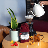 Red velvet drawstring coffee bag, with pour over, potted plant, with pour over, pine apple, strawberries and apricot and class amber coffee mug on wooden coffee table.