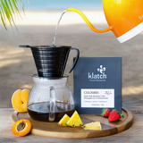 Coffee brewing process with a yellow kettle, coffee filter, and klatch coffee package on a wooden tray with fruits against a beach background.