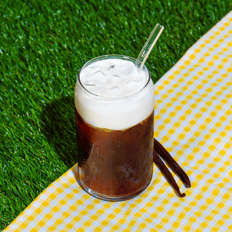 Glass of iced cold brew topped with vanilla foam on a grassy background beside vanilla beans.