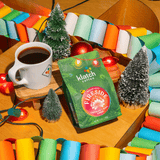 Klatch Coffee Fireside holiday coffee bag displayed beside a mug of coffee, miniature Christmas trees, colorful garlands, and festive lights.