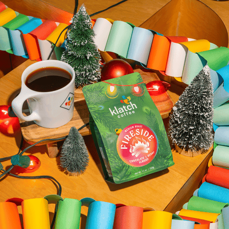 Klatch Coffee Fireside holiday coffee bag displayed beside a mug of coffee, miniature Christmas trees, colorful garlands, and festive lights.