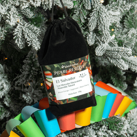 A black drawstring velvet pouch of Klatch Coffee Pearl Reserve El Salvador whole bean coffee hangs on a flocked Christmas tree, with colorful paper garlands visible below.