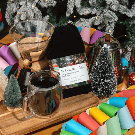 A coffee brewing and serving setup on a wooden tray, featuring a glass pour-over dripper, a glass mug filled with dark coffee, a black drawstring pouch labeled 'Pearl Reserve El Salvador', a small frosted Christmas tree, a metal coffee pot, and colorful paper garlands, with a decorated Christmas tree in the background.