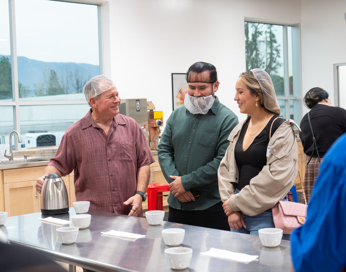 Mike Perry speaking with guests during a Klatch Coffee cupping, with tasting cups and a hot water kettle arranged on the cupping table.
