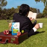 Person sitting on grass reading a book with a briefcase, a chemex of coffee on top with a bag of Out of Office and fruit.