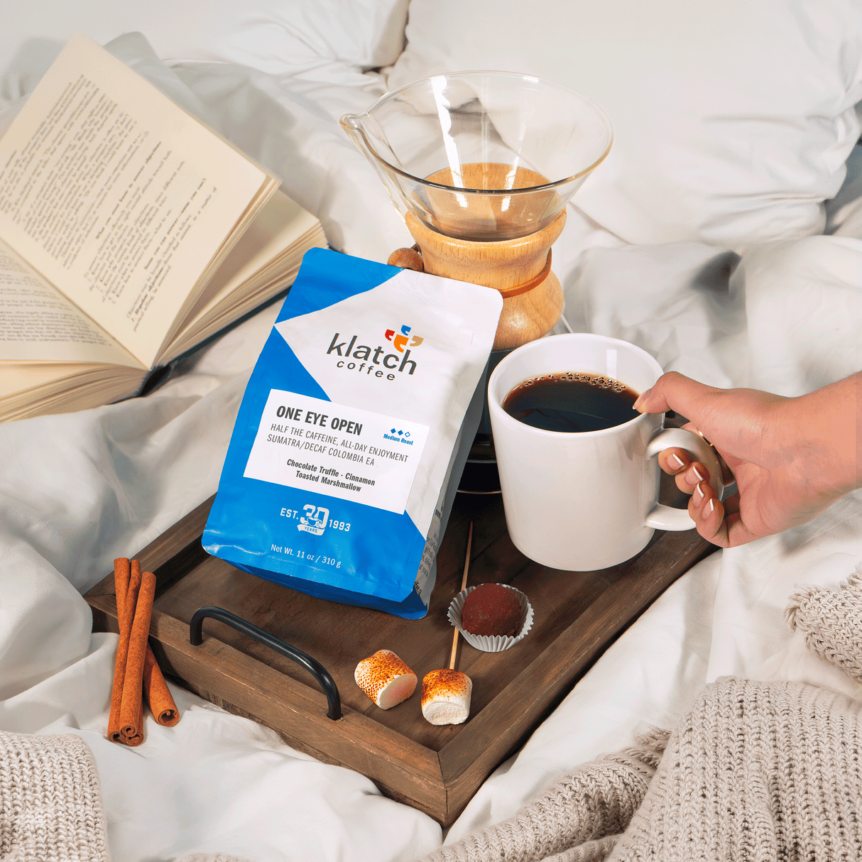 Person holding a cup of coffee next to a package of Klatch coffee on a tray with a book, marshmallow, chocolate truffle and cinnamon. 