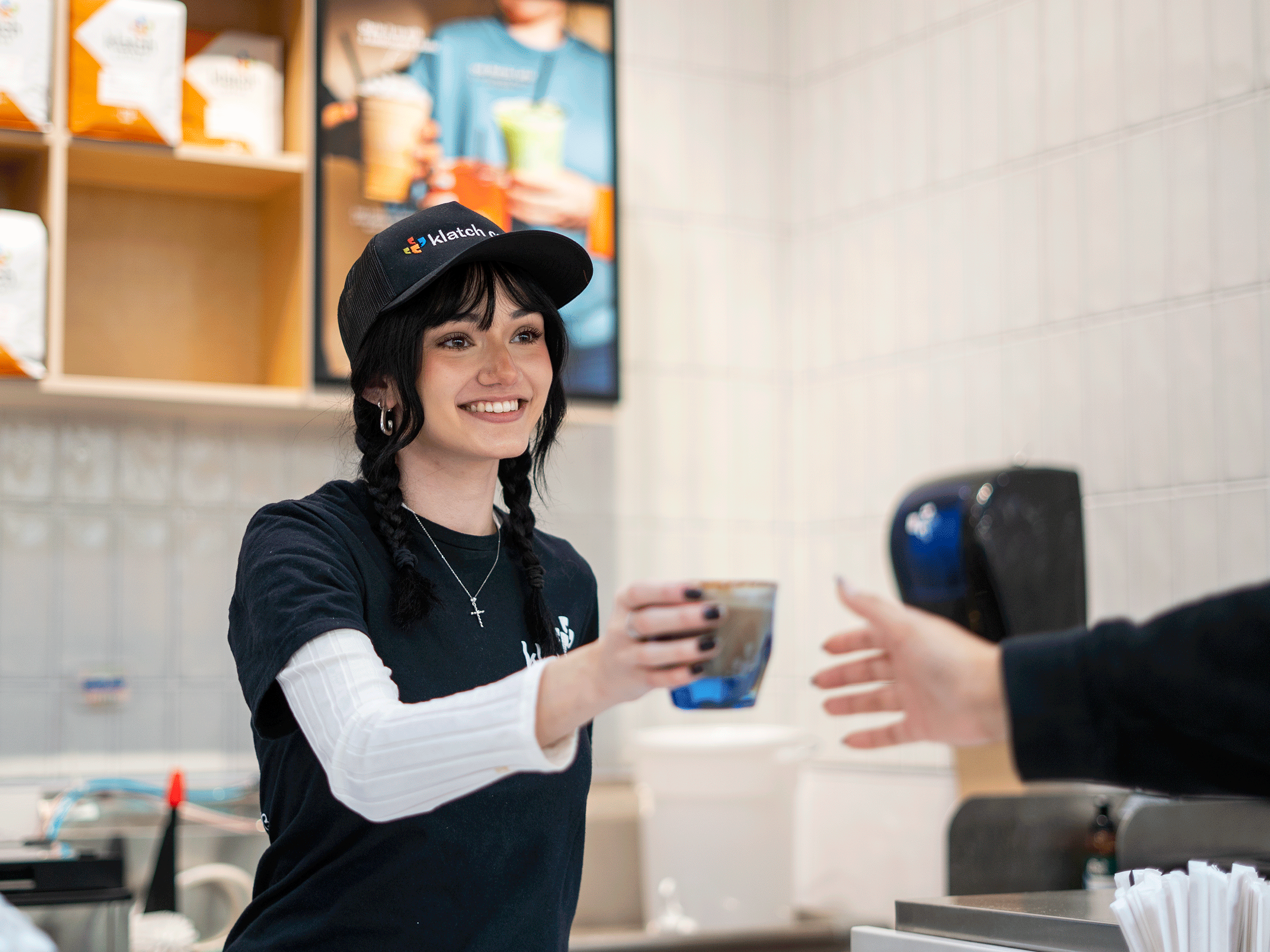 A Klatch Coffee barista hands a drink to a customer across the counter in a café setting.