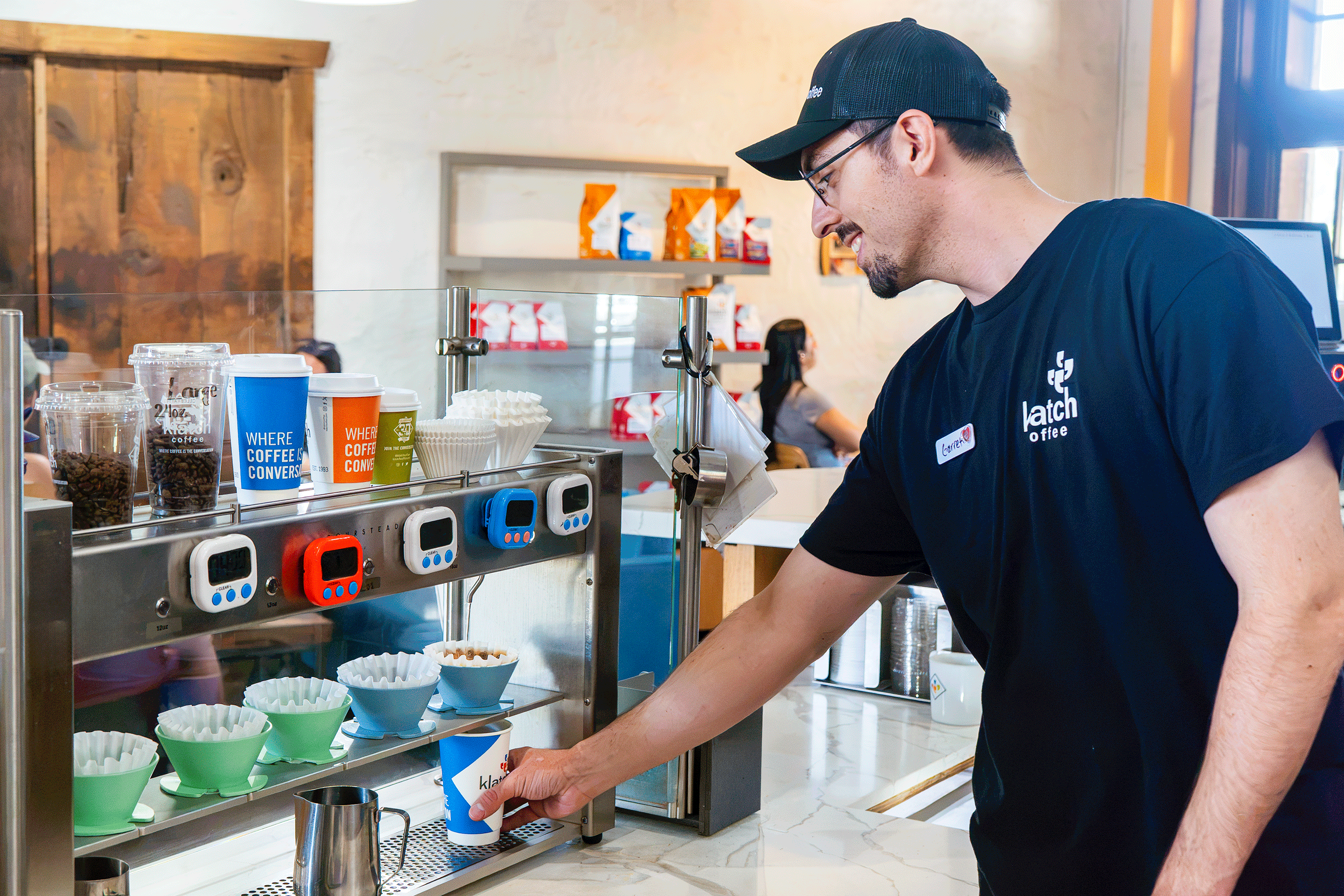 Barista removing a freshly brewed coffee from a Poursteady machine in a café, with colorful brew timers lined up on the brewer.