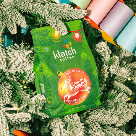 Klatch Coffee Dancing Reindeer holiday coffee bag featuring festive green packaging with a reindeer illustration, nestled among snow-covered Christmas tree branches, twinkling lights, and colorful paper garlands.