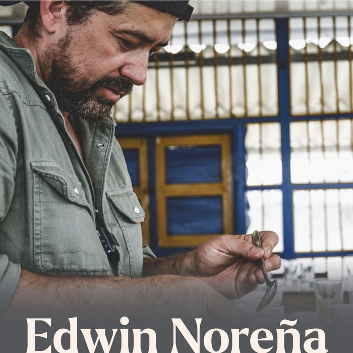 Edwin holding a cupping spoon with 'Edwin Noreña' branding in the foreground, in front of a blue and yellow gate.