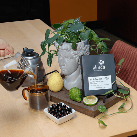A coffee roasting and serving scene on a light wood table, with a hand pouring dark coffee from a glass pour-over pot into a metal mug. A Pearl Reserve El Salvador Pacamara Black Honey on a wooden tray beside a gray bust-shaped planter with trailing green leaves, along with a Asian pear, bergamot, black currant,.