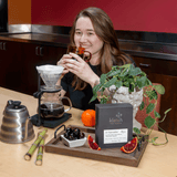 A woman smiles while holding a glass mug in a kitchen setting, with a pour-over coffee setup on a table in front of her. The setup includes a glass carafe, metal kettle, plant, fruit, and a Pearl Reserve box.
