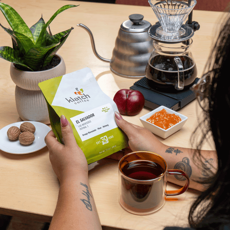 A person holds a bag of Klatch Coffee El Salvador on a wooden table, surrounded by a glass pour-over coffee maker, a metal gooseneck kettle, a mug of brewed coffee, a plum, orange marmalade and nutmage with a small plant.