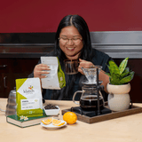 A woman with glasses is smiling while holding a bag of Klatch Coffee Ethiopia and a glass mug, standing behind a coffee bar setup with a pour-over dripper, glass carafe of coffee, oranges, and a potted plant.