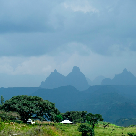 A scenic landscape showing layered blue mountains under a cloudy sky, with green fields, trees, in the foreground.