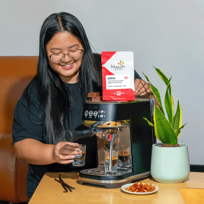 Person preparing espresso on a Breville espresso machine with a bag of Klatch Coffee Eureka blend displayed on top, surrounded by chocolate, vanilla beans, and a potted plant.
