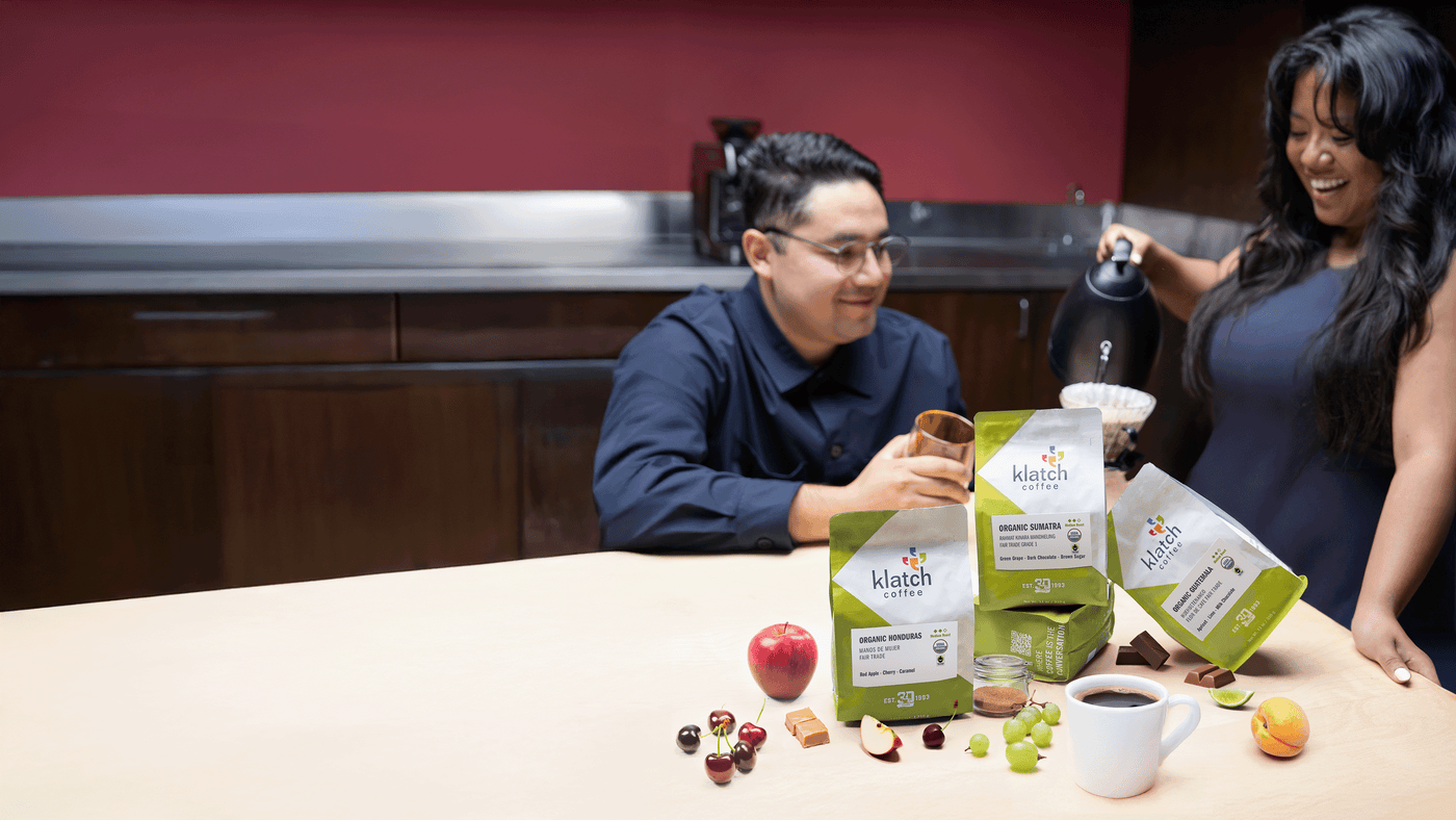 Two people enjoying coffee, one brewing  Klatch Coffee, four green (Fair Trade Coffees) and white coffee bags with coffee mug, fruits on a table.