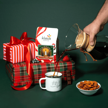 Hand pouring coffee into a mug with a gift, fireside bag of coffee, and bowl of almonds on a green background.