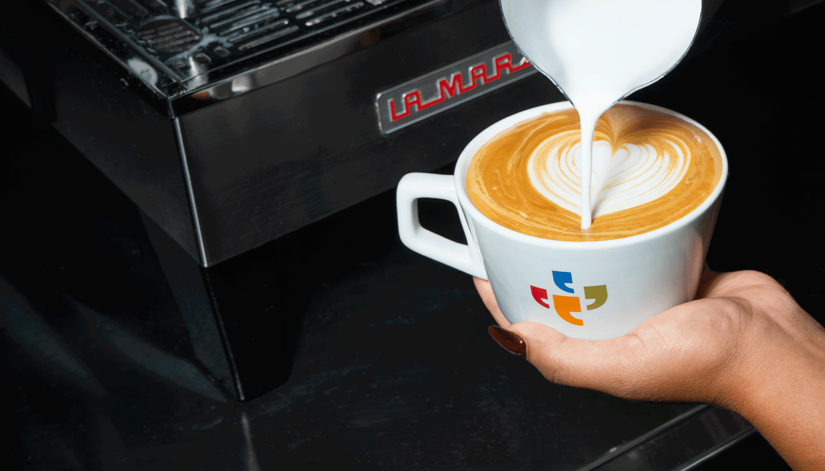 Barista pouring steamed milk into espresso to create latte art beside a La Marzocco espresso machine.