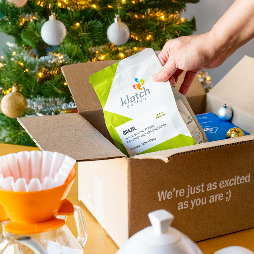 Hand grabbing and holding a Brazil single-origin coffee bag from a shipping box, with a Christmas tree in the background.