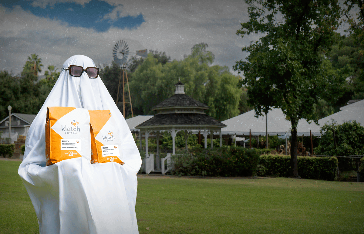A person dressed as a ghost wearing sunglasses holds two orange Klatch Coffee bags on a grassy lawn, with a gazebo and windmill in the background.