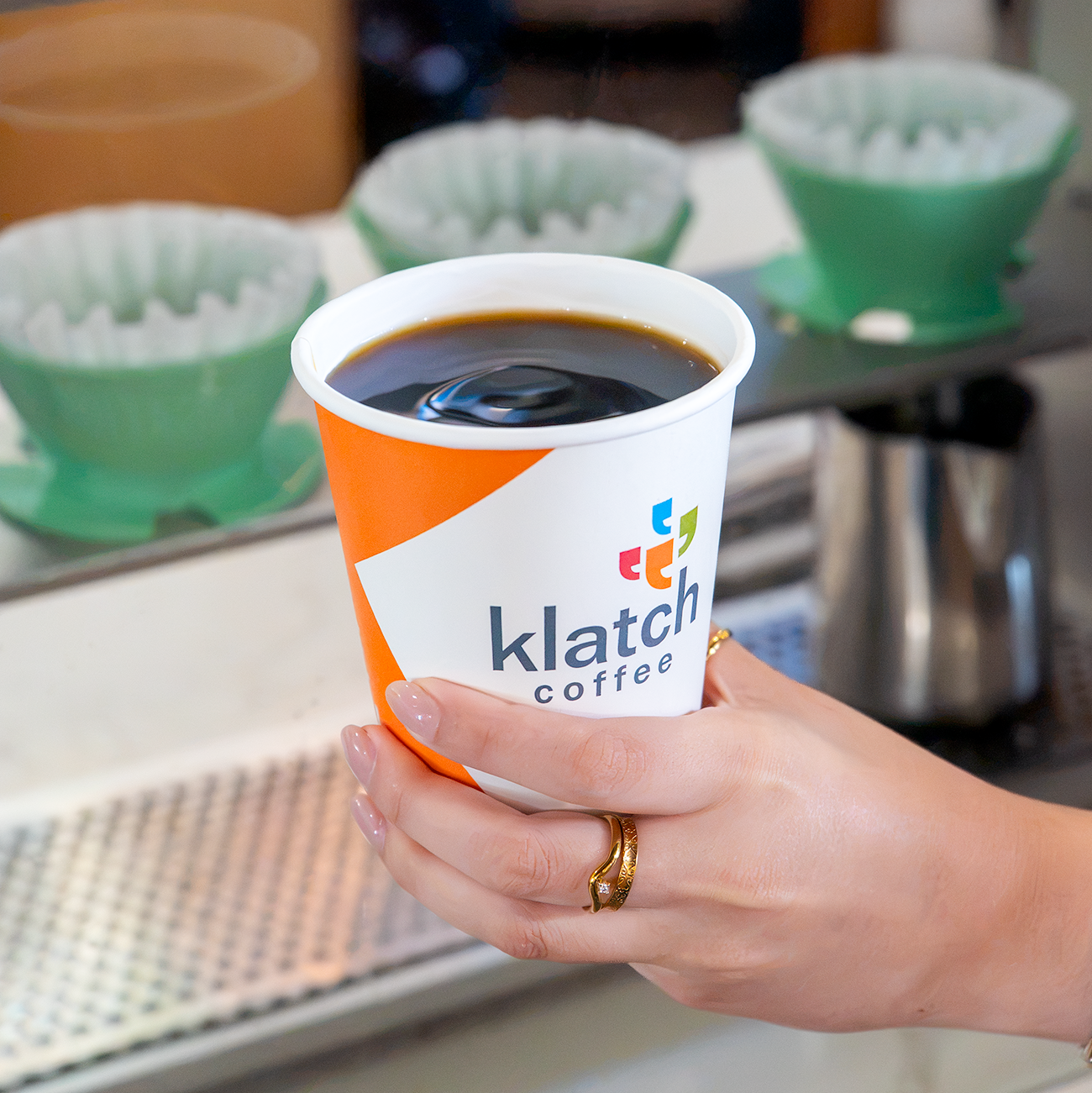 Hand holding an orange Klatch Coffee cup in front of a Poursteady automated pour over brewer.