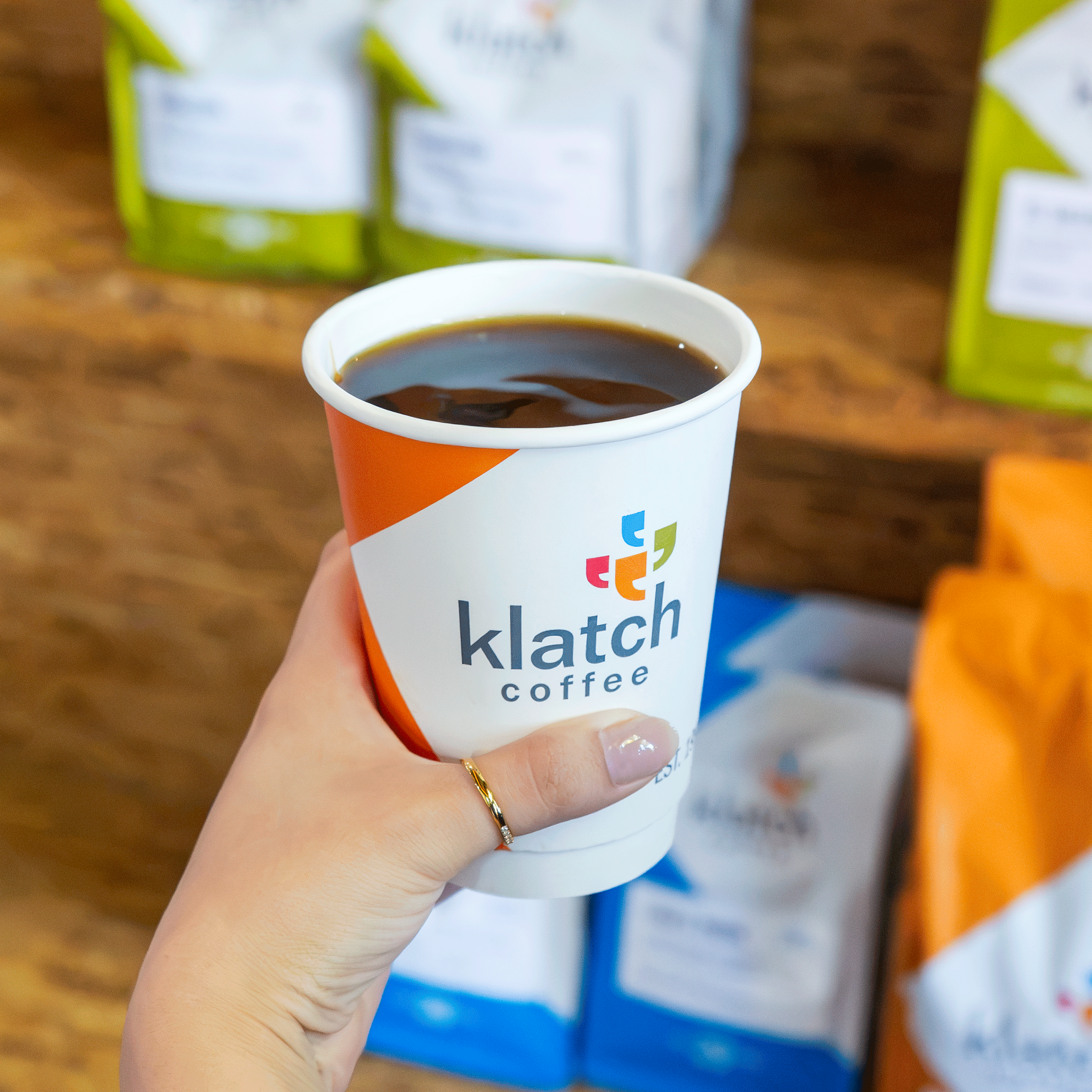 Hand holding an orange Klatch Coffee cup in front of shelves stocked with bags of coffee in a café.