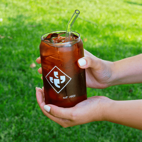 Hands holding a glass of cold brew featuring the Klatch Coffee diamond logo against a grassy background.