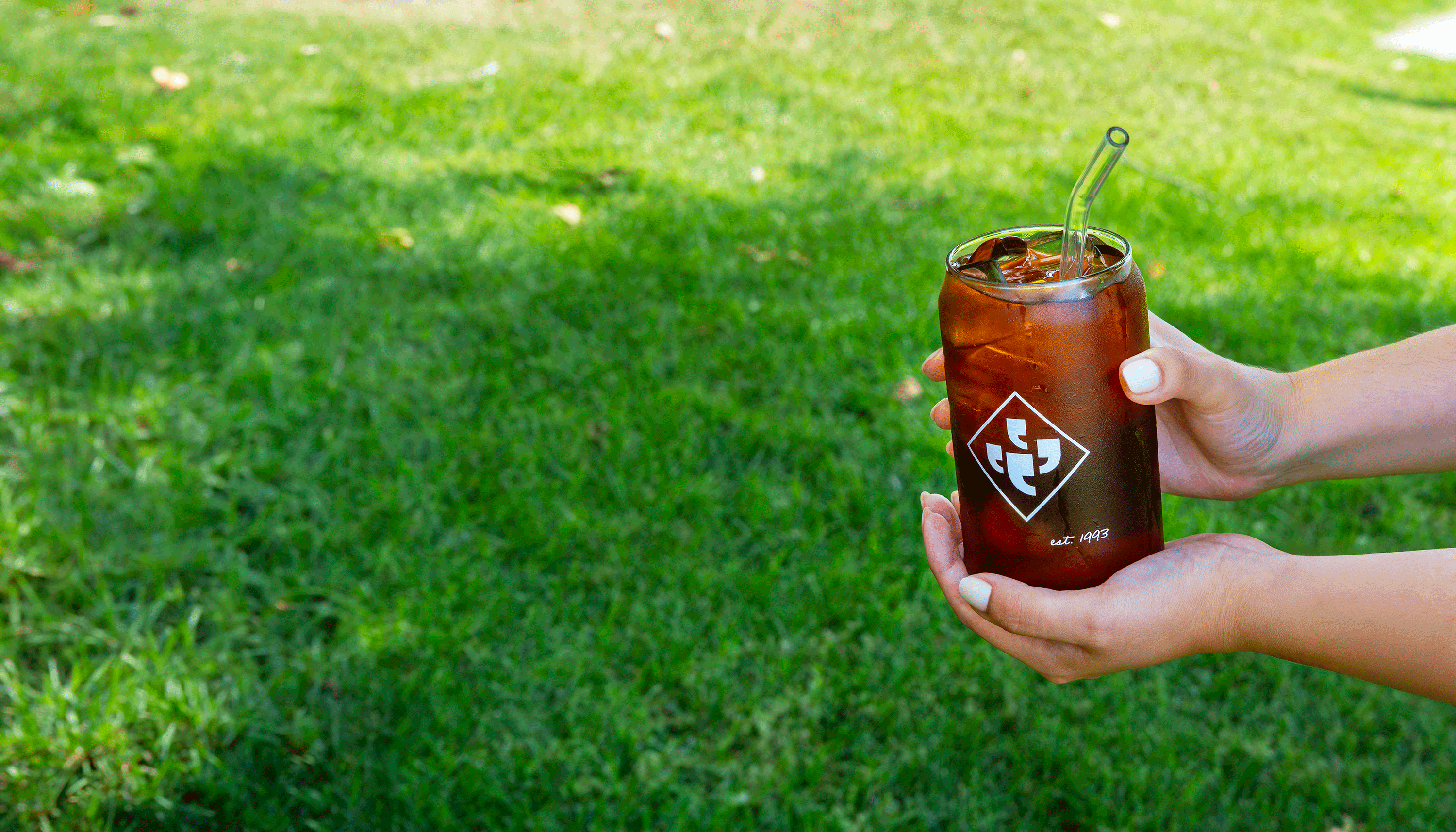 Hands holding a glass of cold brew featuring the Klatch Coffee diamond logo against a grassy background.
