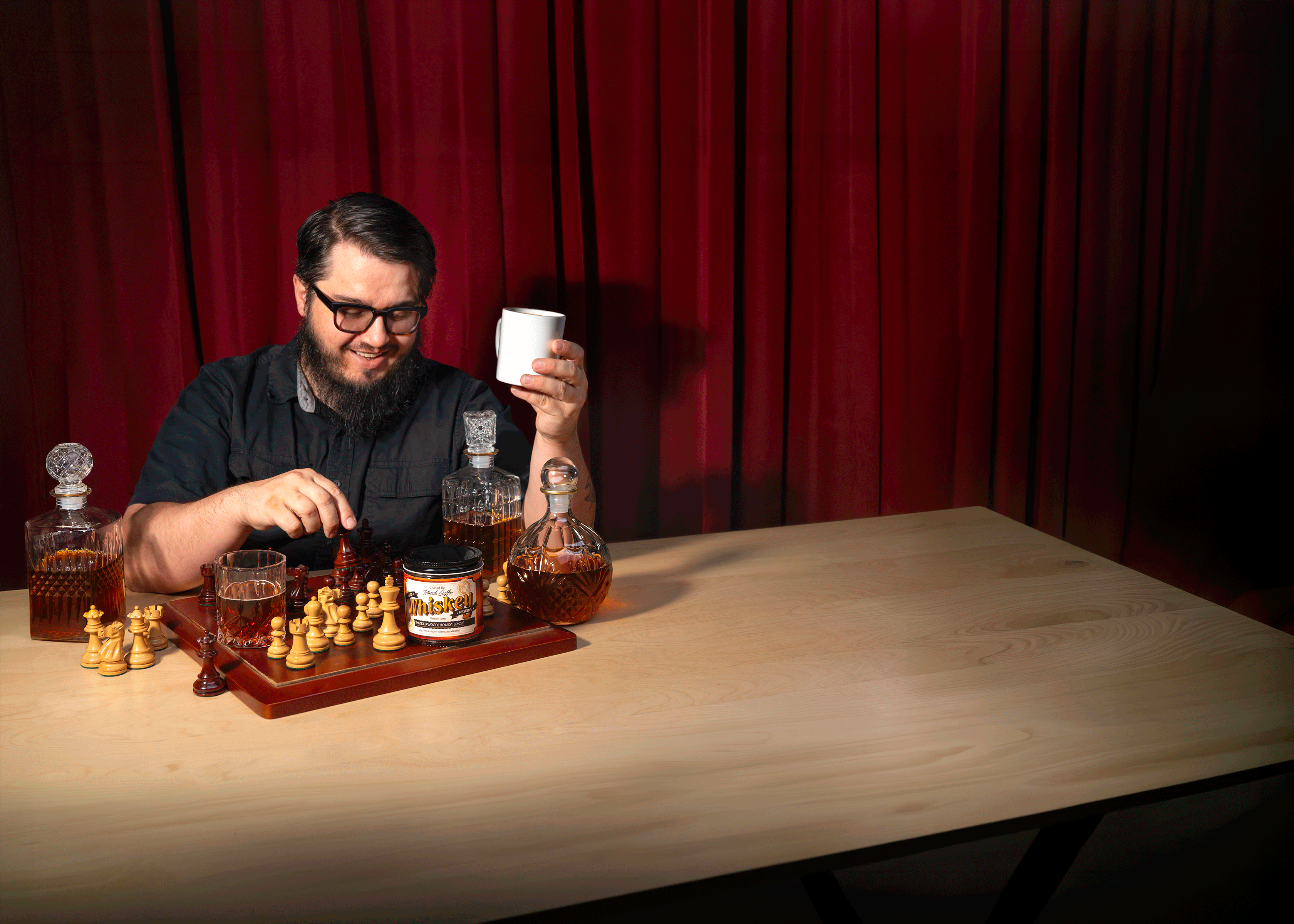 A man wearing glasses plays chess while holding a coffee mug. On the chessboard sits a bag of Klatch Coffee Aged Whiskey Reserve coffee, surrounded by whiskey decanters.