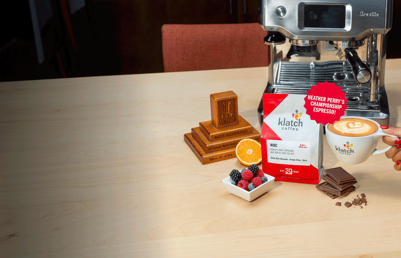 Klatch Coffee's WBC espresso bag on a table with chocolate, orange, and berries. An espresso machine, WBC trophy, latte, and a graphic reading "Heather Perry's Championship Espresso" appear in the background.