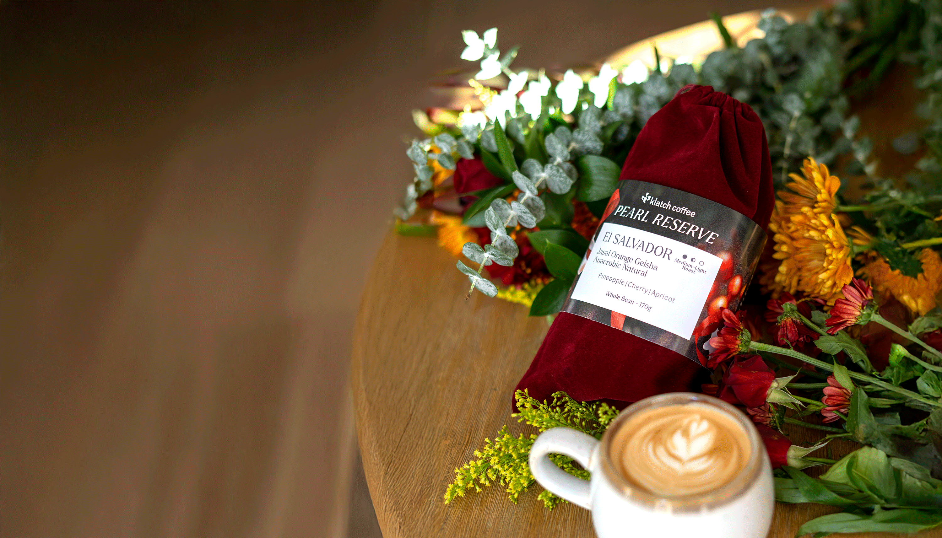 El Salvador Orange Geisha in a red velvet bag on a table, surrounded by flowers and a latte.