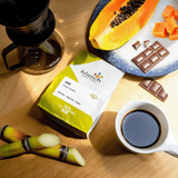Java Gunung coffee bag on wooden table, glass ethoz pour-over with white mug with coffee. Fruit of flavor notes of chocolate bar and  half cut papaya  on a plate with two stacks of sugarcane laid on table at the bottom of the bag..