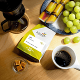 Klatch Coffee Java Gunung Tikukur bag with a cup of coffee, grapes, sugarcane, small glass bowl of caramel cubes and a brewing device with a mug of coffee on a wooden table.