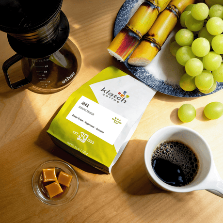 Klatch Coffee Java Gunung Tikukur bag with a cup of coffee, grapes, sugarcane, small glass bowl of caramel cubes and a brewing device with a mug of coffee on a wooden table.