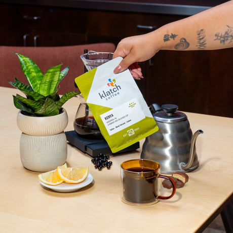A hand holds a bag of Klatch Coffee Kenya Kaganda AA in front of a coffee setup on a light wood table, including a gooseneck kettle, brewed coffee in a glass mug, lemon slices, and a potted plant.