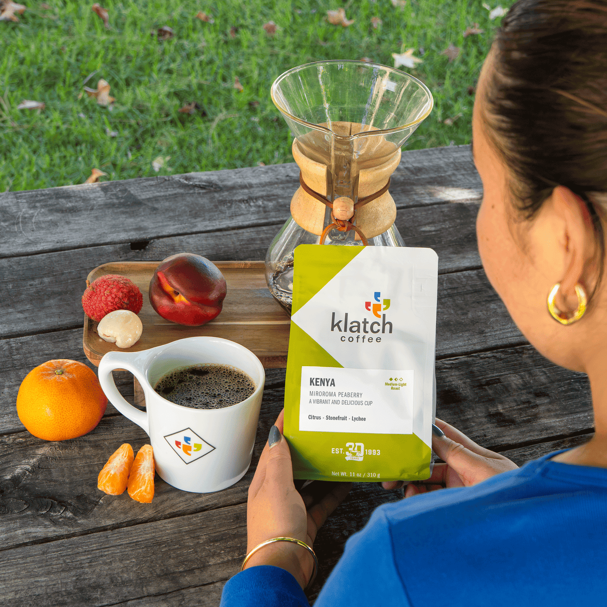 A person outdoors on a wooden table holds a bag of Klatch Coffee Kenya, with a glass pour-over coffee maker, a cup of black coffee, and fruit nearby.
