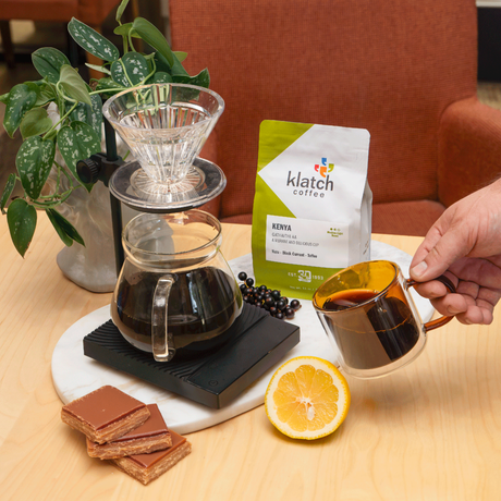 A hand holds a glass mug filled with brewed coffee next to a bag of Klatch Coffee Kenya on a light wood table. The scene includes a pour-over coffee maker, a glass carafe, a sliced yuzu, toffee, and black currant with a potted plant in the background.
