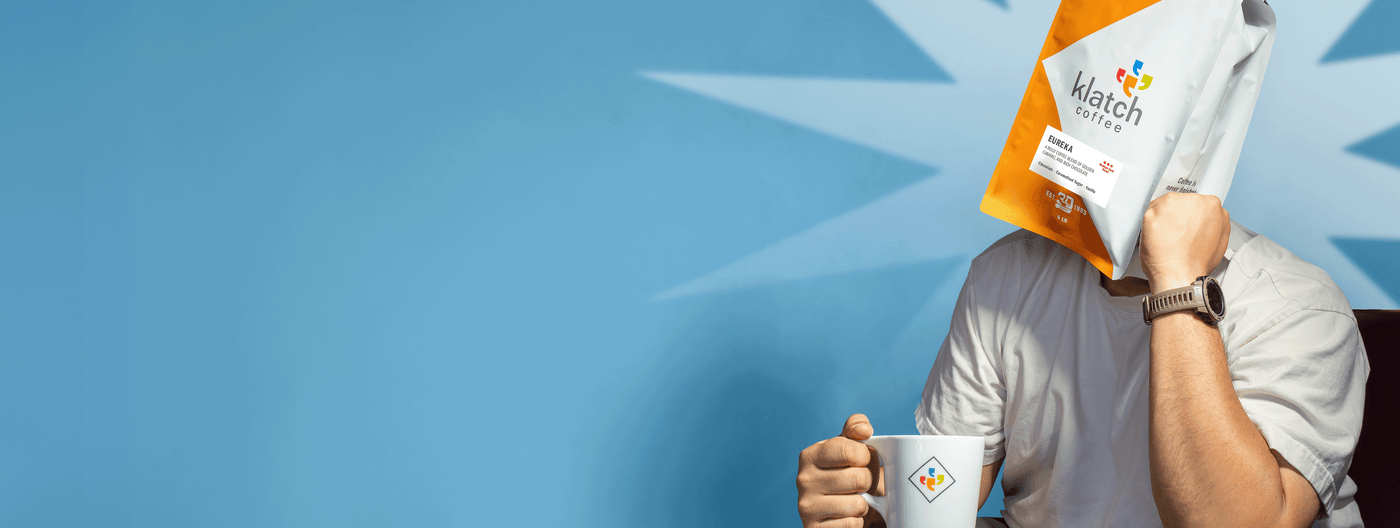Man holding a coffee cup and coffee bag on head against a blue background.