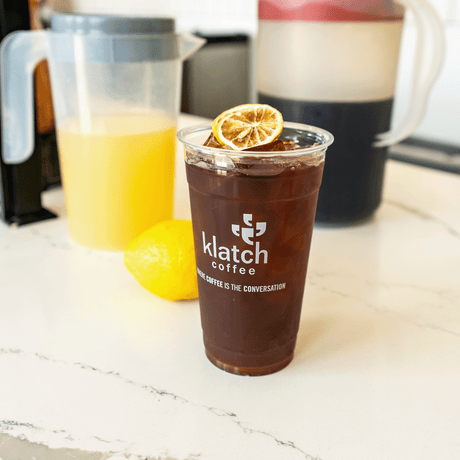 pitcher of Lemonade and Cold Brew in background on counter with plastic cup of Lemonade Cold Brew with dried lemon on top of ice.