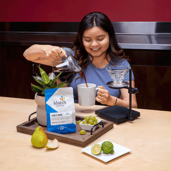 Woman pouring freshly brewed coffee from a glass carafe into a white mug beside a pour over dripper. A bag of Klatch Coffee Lion's Pride blend is displayed with fresh pear, white grapes, and bergamot to highlight the coffee's tasting notes.
