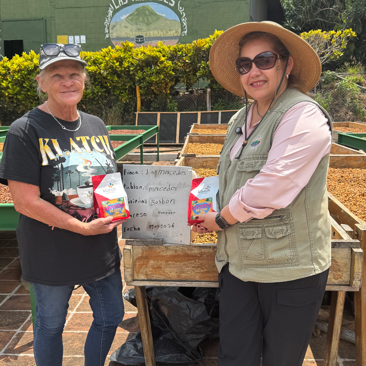 Luci and Cindy infront of drying beds holding Mujeres De Cafe bags.