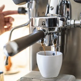 A close-up of a brushed stainless steel LELIT MaraX3 Espresso Machine dispensing dark coffee into a small white ceramic cup. A hand is visible on the left operating the machine.