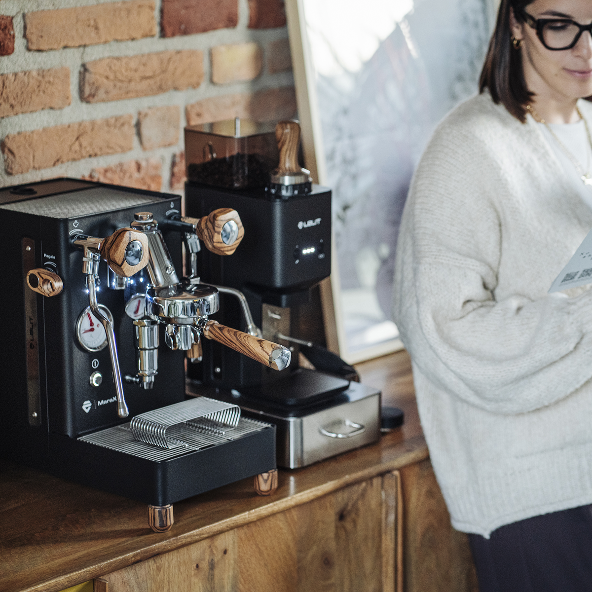A black LELIT MaraX3 Espresso Machine with wooden handles and a matching coffee grinder sit on a wooden counter in a cafe setting, with a woman in a white sweater standing beside them.