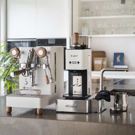 A white LELIT MaraX3 Espresso Machine set with a matching William Coffee Grinder and accessories, arranged on a kitchen countertop in a bright, modern kitchen.