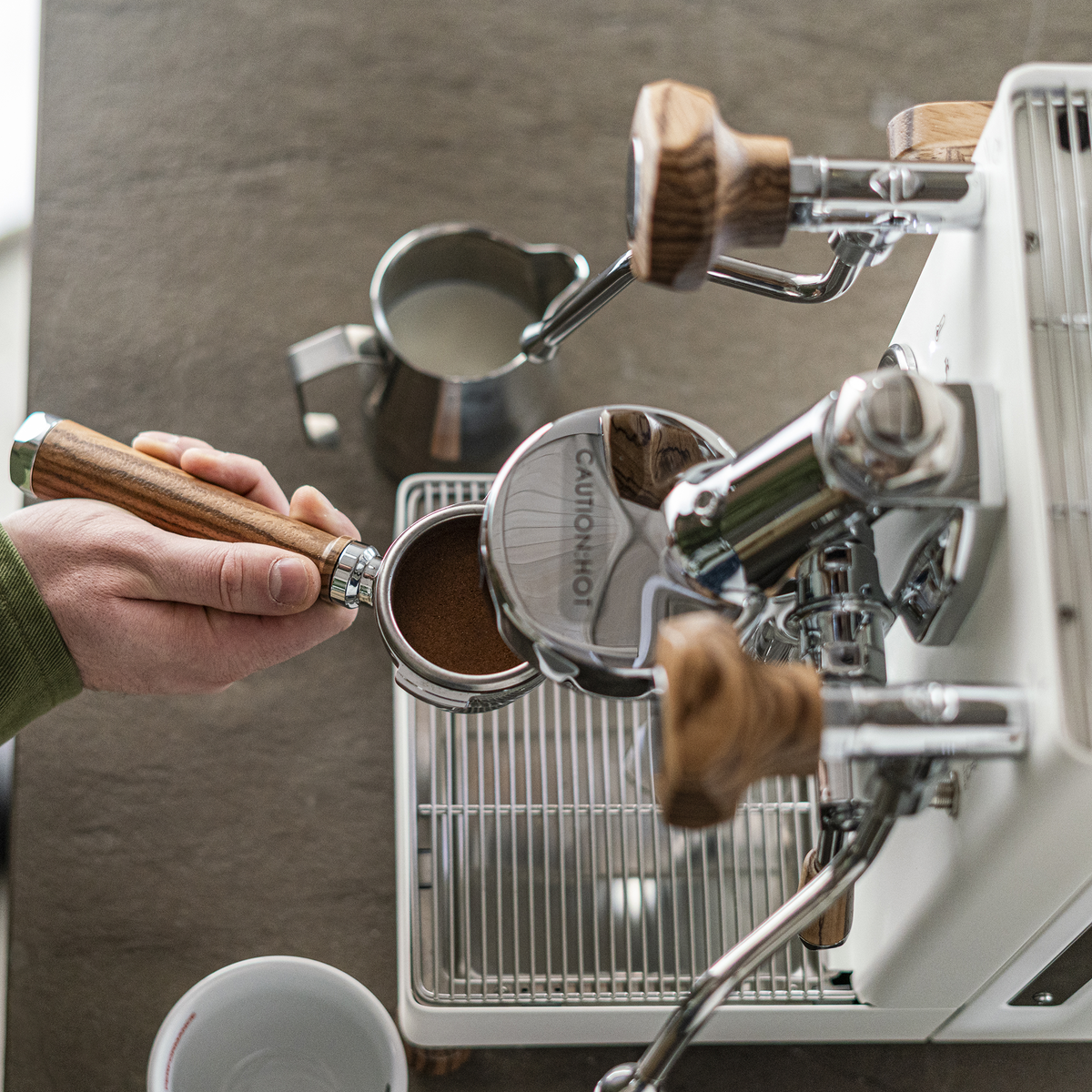 A close-up of a hand holding a portafilter with espresso powder over a white and chrome espresso machine, with a milk pitcher and coffee cup nearby on a gray countertop.