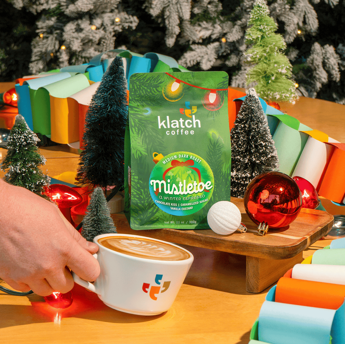 Klatch Coffee Mistletoe Winter Espresso holiday coffee bag displayed among miniature Christmas trees, colorful paper garlands, glowing holiday lights, and festive ornaments, with a hand holding a white Klatch Coffee latte art cup in the foreground.