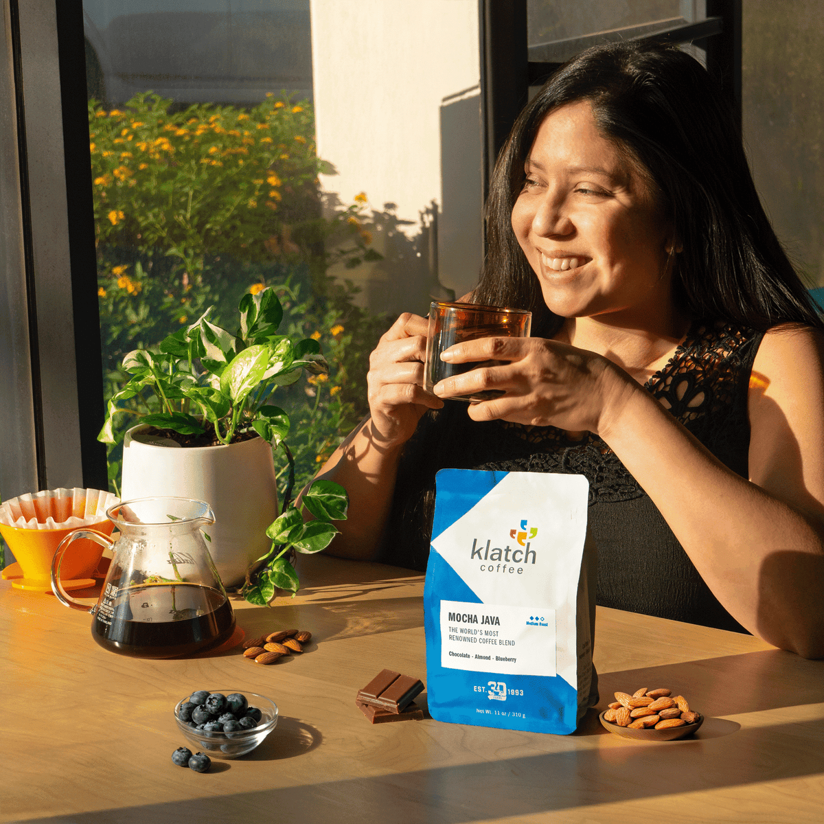 Woman enjoying a cup of coffee with a Klatch coffee bag on a table by a window with plants, almonds, blueberries and chocolate bar on table.