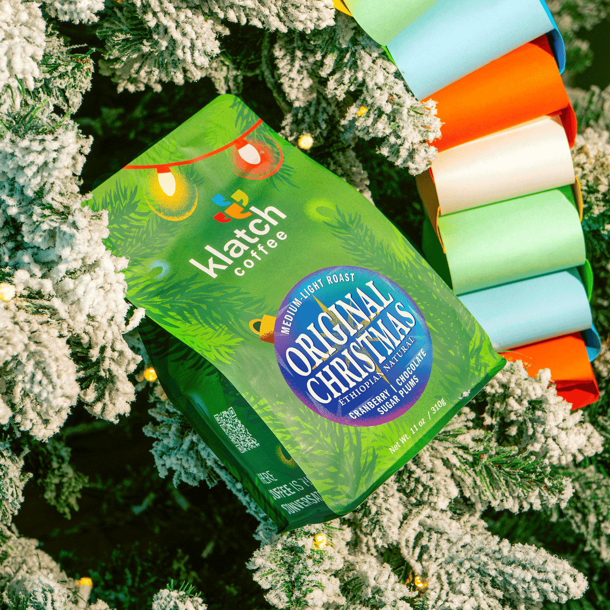 Klatch Coffee Original Christmas holiday coffee bag featuring festive green packaging with colorful Christmas lights, nestled among snow-covered Christmas tree branches, twinkling lights, and a vibrant multicolored paper garland.
