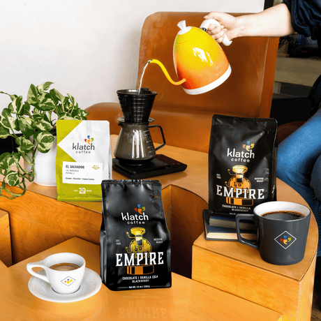 Person pouring hot water into brewing device on coffee table with three coffee bags, coffee mug and espresso.